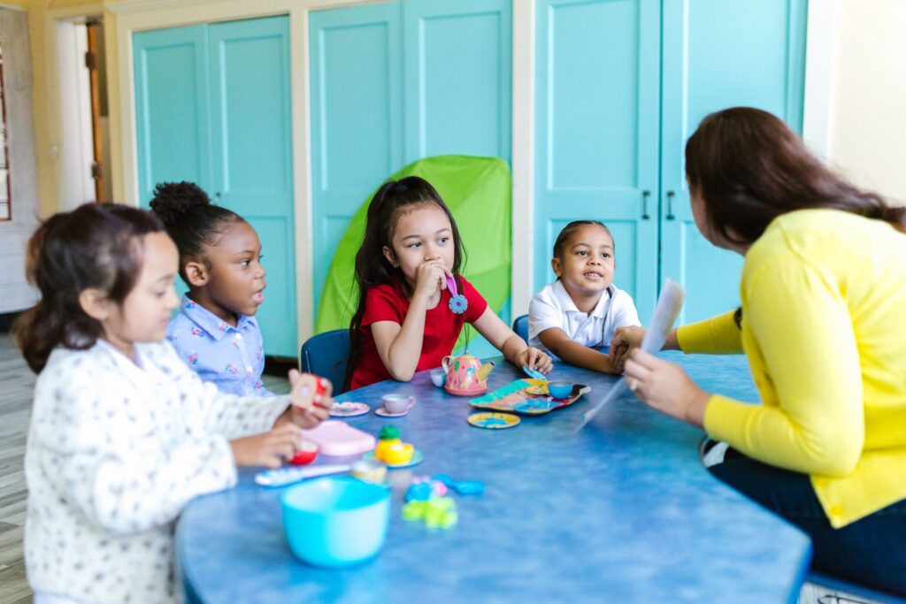 Four young children seated at a table with a toy tea set and a teacher in front of them